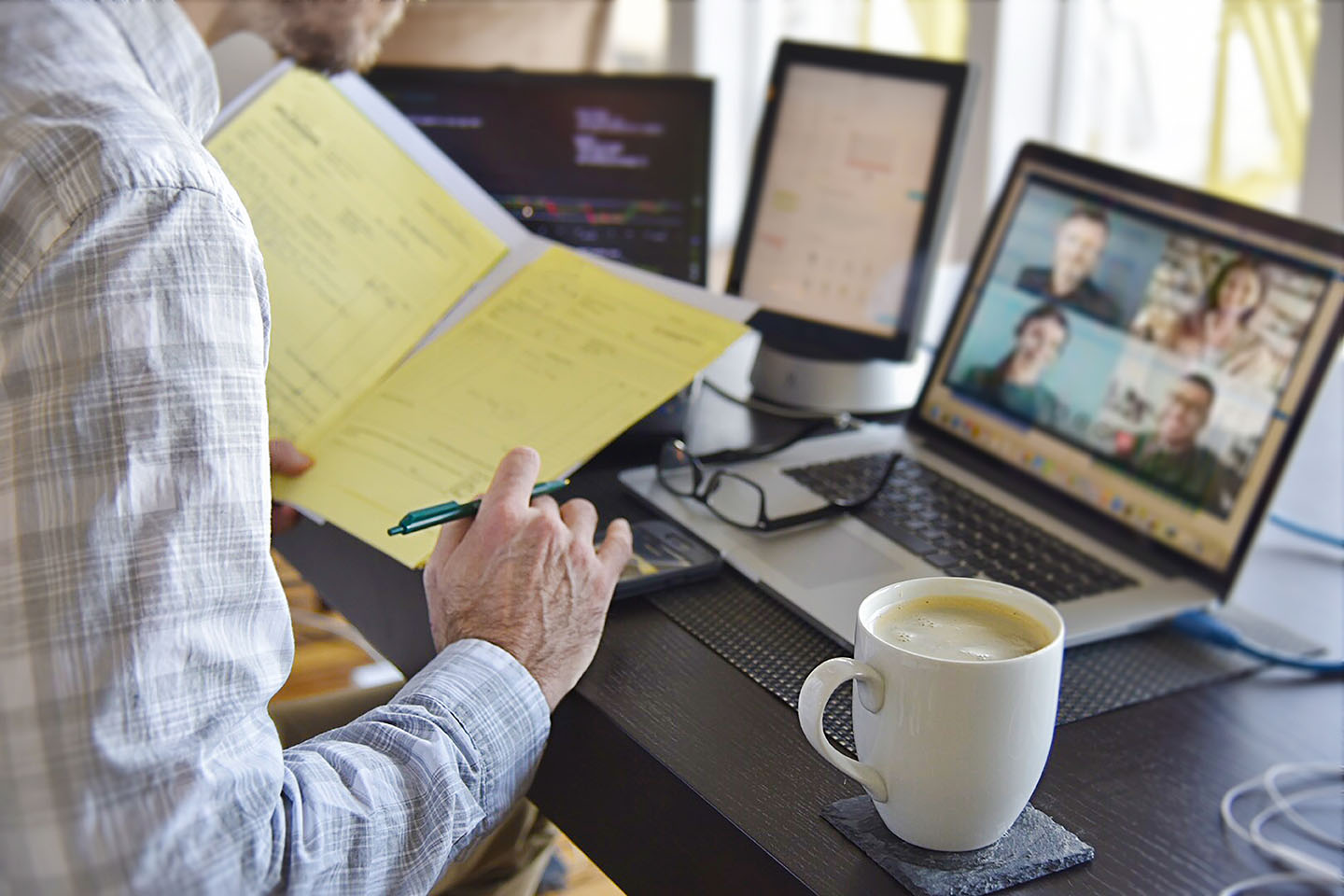 Man working from home signing contract documents while in a virtual video chat meeting on computer screens.